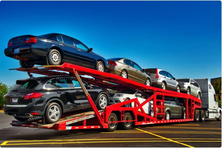Red car carrier trailer transporting multiple vehicles on a clear day in a parking lot.